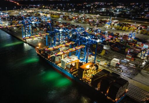 Aerial view of a large Maersk container ship docked at a busy port terminal at night with illuminated gantry cranes and stacks of shipping containers in Ghana. Aerial view of a large Maersk container ship docked at a busy port terminal at night with illuminated gantry cranes and stacks of shipping containers in Ghana.