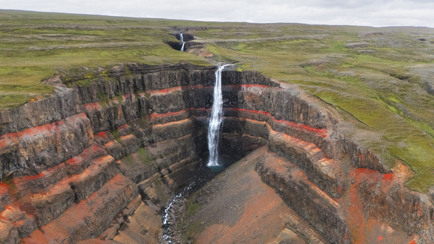 Aerial view of Hengifoss waterfall plunging down the cliff face cutting through layers of vibrant red clay contrasting with the green landscape Hengifoss Iceland.