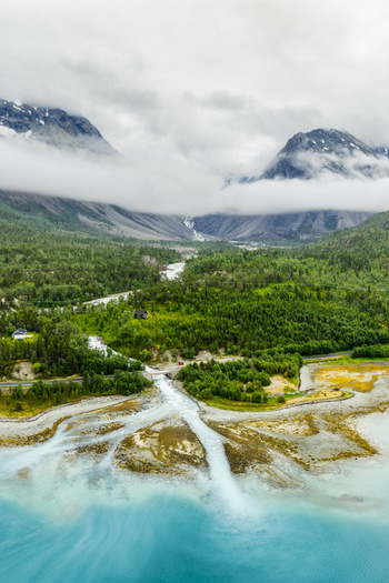 Aerial view of the glacial river in Faueldalen flowing into the turquoise fjord surrounded by lush green forests and dramatic mountains in Jovik Troms Norway.