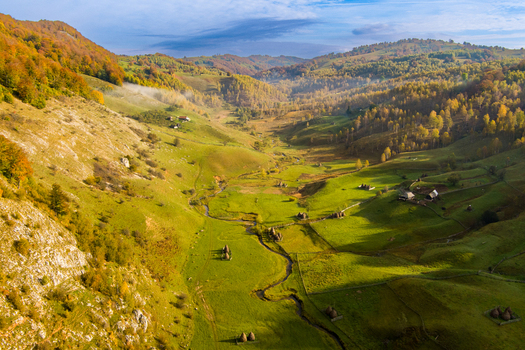 Aerial view of Fundatura Ponorului featuring a lush green valley with a winding stream traditional haystacks and autumn colored hills Ohaba Ponor Hunedoara Romania.