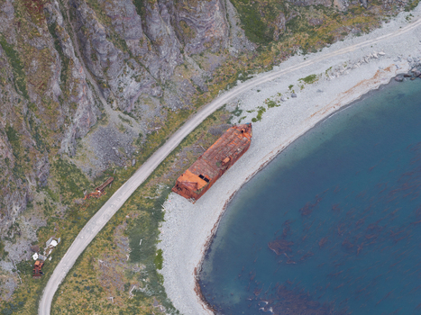 Aerial view of a rusted shipwreck resting on a rocky beach next to a gravel road and steep cliffs in Unalaska Alaska United States.