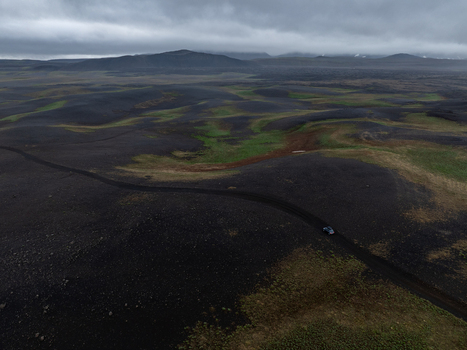 Aerial view of a car driving along a winding dirt road through a vast dark volcanic landscape with patches of green moss under an overcast sky in Iceland. Aerial view of a car driving along a winding dirt road through a vast dark volcanic landscape with patches of green moss under an overcast sky in Iceland.