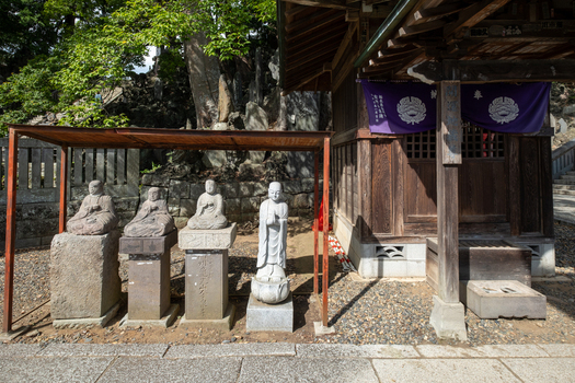 View of aged stone statues stand serenely beside a weathered wooden structure adorned with purple banners blending history with nature Narita Chiba Japan. View of aged stone statues stand serenely beside a weathered wooden structure adorned with purple banners blending history with nature Narita Chiba Japan.