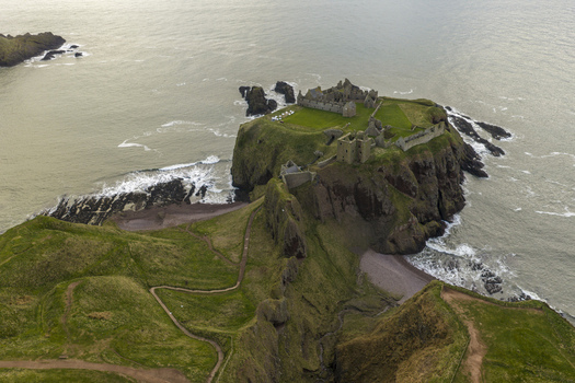 Aerial view of Dunnottar Castle ruins perched on a dramatic rocky headland surrounded by the North Sea and green cliffs in Stonehaven Scotland United Kingdom. Aerial view of Dunnottar Castle ruins perched on a dramatic rocky headland surrounded by the North Sea and green cliffs in Stonehaven Scotland United Kingdom.