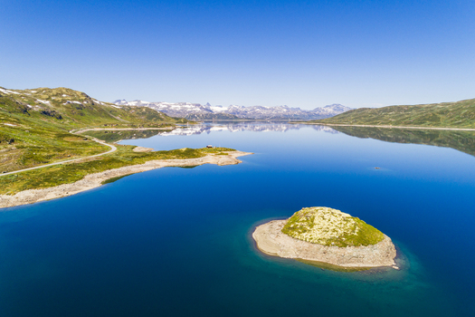 Aerial view of the calm blue waters of Lake Tyin with a small grassy island surrounded by rugged mountains and a winding road in Tyinkrysset Innlandet Norway.