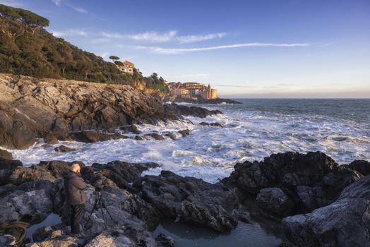 View of the historic village of Tellaro perched on the rocky coastline with waves crashing against the shore and a photographer on the rocks Tellaro Liguria Italy. View of the historic village of Tellaro perched on the rocky coastline with waves crashing against the shore and a photographer on the rocks Tellaro Liguria Italy.