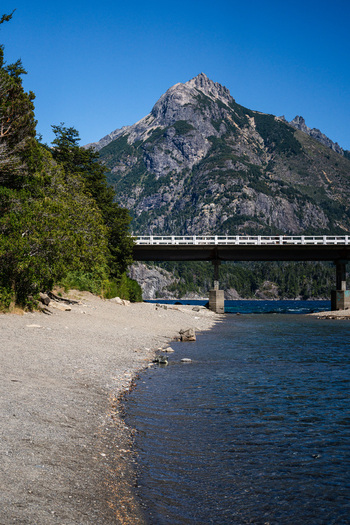 View of Puente Arroyo La Angostura spanning the water with a rocky shoreline and a large mountain in the background under a clear blue sky in San Carlos de Bariloche Rio Negro Argentina.