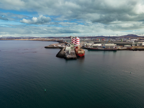 Hafnarfjordur Iceland 17 April 2026 Aerial view of the port featuring a red and white striped silo a cargo ship docked at the pier and snow capped mountains under a cloudy sky. Hafnarfjordur Iceland 17 April 2026 Aerial view of the port featuring a red and white striped silo a cargo ship docked at the pier and snow capped mountains under a cloudy sky.