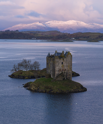 Aerial view of Castle Stalker on a small tidal islet surrounded by calm water with snow capped mountains under a soft sky in Appin Scotland United Kingdom. Aerial view of Castle Stalker on a small tidal islet surrounded by calm water with snow capped mountains under a soft sky in Appin Scotland United Kingdom.