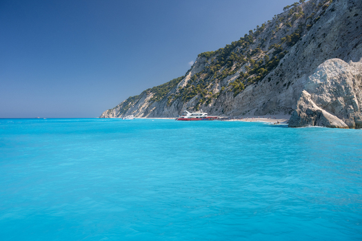 View of the turquoise waters of the Ionian Sea at a beach with steep white cliffs and a passenger boat docked near the shore in Lefkada Greece.