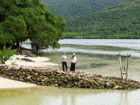 Aerial view of two figures stand on a rocky outcrop framed by verdant trees against the tranquil waters of Bobby Beach Karimunjawa Central Java Indonesia.