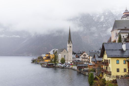 View of the historic village of Hallstatt featuring the iconic church spire and alpine houses along the lake under a misty mountain backdrop in Hallstatt Upper Austria Austria. View of the historic village of Hallstatt featuring the iconic church spire and alpine houses along the lake under a misty mountain backdrop in Hallstatt Upper Austria Austria.