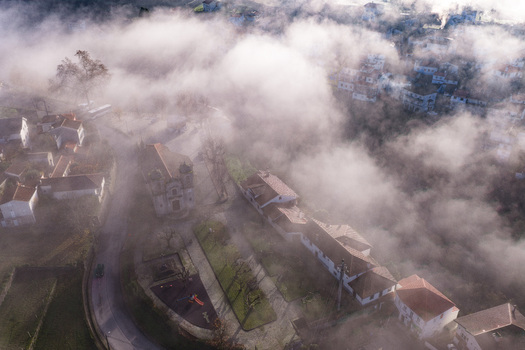 Aerial view of Avo village with its historic church and red tiled houses partially shrouded in thick morning mist and fog Oliveira do Hospital Coimbra District Portugal.