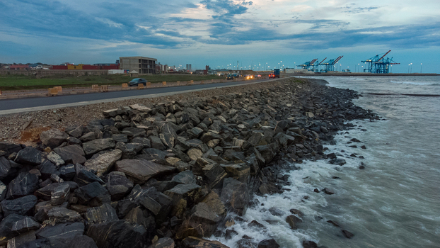 Aerial view of the Accra Tema Beach Road with large rocks forming a breakwater shipping cranes in the distance and a cloudy sky at dusk in Accra Greater Accra Region Ghana.