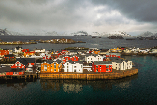 Aerial view of vibrant red yellow and white buildings clustered at the edge of the turquoise sea against a backdrop of snow dusted islands Henningsv&aelig;r Nordland Norway.
