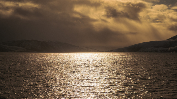 View of the serene waters reflecting a golden sky under brooding clouds with distant hills framing the horizon Brensholmen Troms Norway. View of the serene waters reflecting a golden sky under brooding clouds with distant hills framing the horizon Brensholmen Troms Norway.
