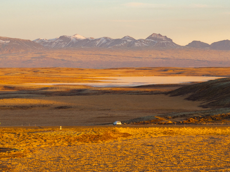 Aerial view of a vast golden landscape with a road a small car and snow capped mountains in the distance in Reykjanesbaer Iceland. Aerial view of a vast golden landscape with a road a small car and snow capped mountains in the distance in Reykjanesbaer Iceland.