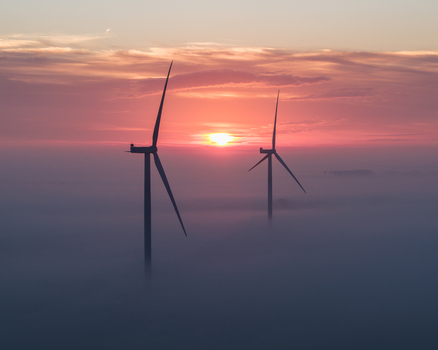 Aerial view of wind turbines emerging from a thick layer of morning fog under a vibrant orange sunrise sky in Vrouwenpolder Zeeland Netherlands.