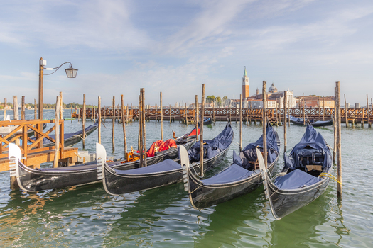 View of traditional black gondolas moored to wooden poles in the calm water of the lagoon with historic Venetian architecture in the background in Venice Veneto Italy. View of traditional black gondolas moored to wooden poles in the calm water of the lagoon with historic Venetian architecture in the background in Venice Veneto Italy.