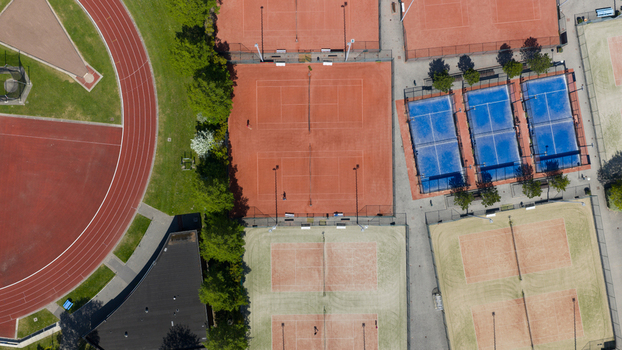 Aerial view of Tennis Padel Club Heiloo featuring red clay and blue padel courts next to an athletic running track and lush green trees in Heiloo Noord Holland Netherlands.