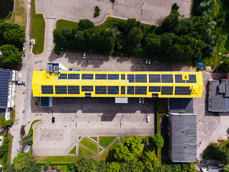 Aerial view of a long yellow industrial building featuring extensive rooftop solar panels surrounded by a parking lot and green trees in Valdlauci Kekava Municipality Latvia. Aerial view of a long yellow industrial building featuring extensive rooftop solar panels surrounded by a parking lot and green trees in Valdlauci Kekava Municipality Latvia.