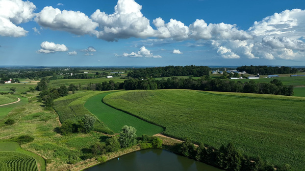Aerial view of Kilmer Road farmland with lush green cornfields a small pond and dense tree lines under a bright blue sky in Rapho Township Pennsylvania United States. Aerial view of Kilmer Road farmland with lush green cornfields a small pond and dense tree lines under a bright blue sky in Rapho Township Pennsylvania United States.