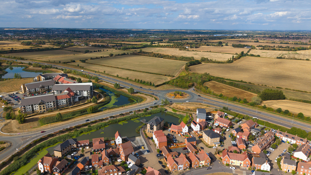Aerial view of a modern residential housing estate featuring red brick houses a small lake and a nearby roundabout with surrounding agricultural fields in Bedford England United Kingdom. Aerial view of a modern residential housing estate featuring red brick houses a small lake and a nearby roundabout with surrounding agricultural fields in Bedford England United Kingdom.