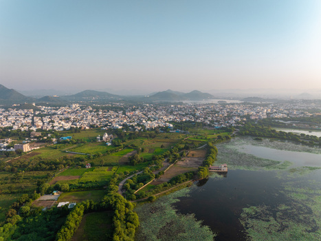 Aerial view of the cityscape of Udaipur with white buildings surrounding green fields and a large lake with vegetation under a clear sky in Udaipur Rajasthan India.