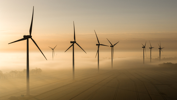 Aerial view of wind turbines piercing through the ethereal fog casting long shadows across the fields in the golden light of dawn Wislina Pomeranian Voivodeship Poland. Aerial view of wind turbines piercing through the ethereal fog casting long shadows across the fields in the golden light of dawn Wislina Pomeranian Voivodeship Poland.