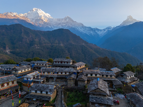 Aerial view of a traditional stone village with slate roofs and colorful prayer flags nestled in the Himalayan mountains under a clear sky in Nepal.