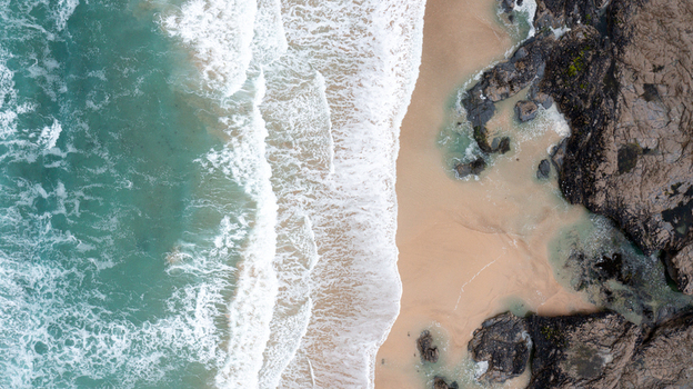 Aerial view of turquoise waves crashing onto the sandy shore and dark coastal rocks at Constantine Bay Padstow Cornwall United Kingdom. Aerial view of turquoise waves crashing onto the sandy shore and dark coastal rocks at Constantine Bay Padstow Cornwall United Kingdom.
