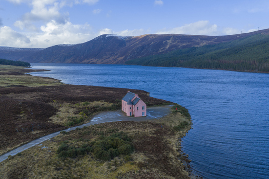 Aerial view of The Pink House on the shore of Loch Glass surrounded by rugged mountains and blue water in Dingwall Scotland United Kingdom. Aerial view of The Pink House on the shore of Loch Glass surrounded by rugged mountains and blue water in Dingwall Scotland United Kingdom.