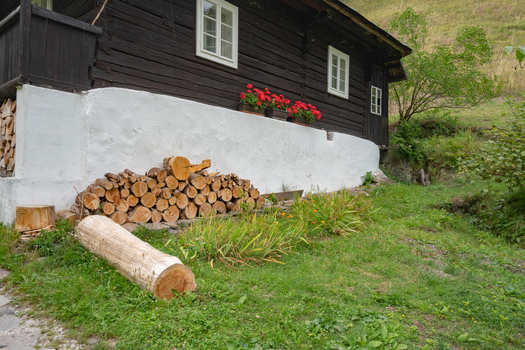 View of a quaint dark wood cabin with white trim and vibrant red flowers sits atop a white stone base firewood stacked nearby Prasnica Banska Bystrica Region Slovakia. View of a quaint dark wood cabin with white trim and vibrant red flowers sits atop a white stone base firewood stacked nearby Prasnica Banska Bystrica Region Slovakia.