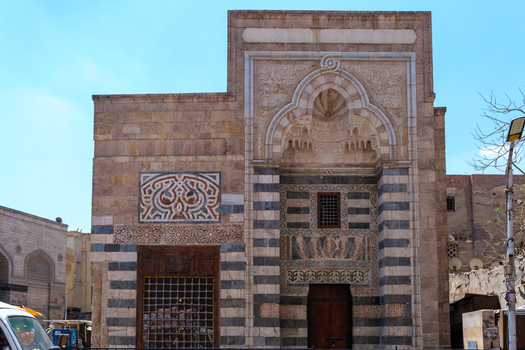 View of the Zawya and Sabil of Sultan Faraj Ibn Barquq featuring intricate stone carvings ablaq striped masonry and a decorative portal under a clear blue sky in Cairo Egypt. View of the Zawya and Sabil of Sultan Faraj Ibn Barquq featuring intricate stone carvings ablaq striped masonry and a decorative portal under a clear blue sky in Cairo Egypt.