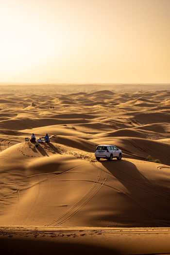 View of Erg Chebbi sand dunes at golden hour with a parked SUV and people resting on the sand in Errachidia Province Draa Tafilalet Morocco.