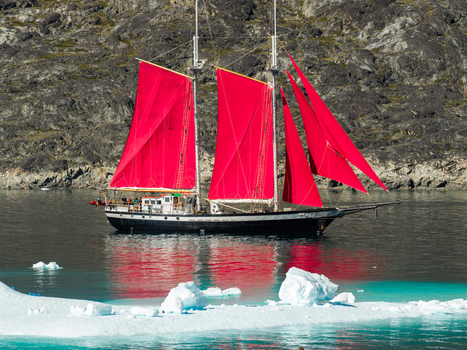 Aerial view of a schooner with vibrant red sails navigating through icy waters near a rocky coastline in Greenland. Aerial view of a schooner with vibrant red sails navigating through icy waters near a rocky coastline in Greenland.