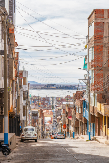 Puno Peru 23 June 2026 View of a steep street with brick buildings overhead power lines and a vintage car leading down towards the distant Lake Titicaca.