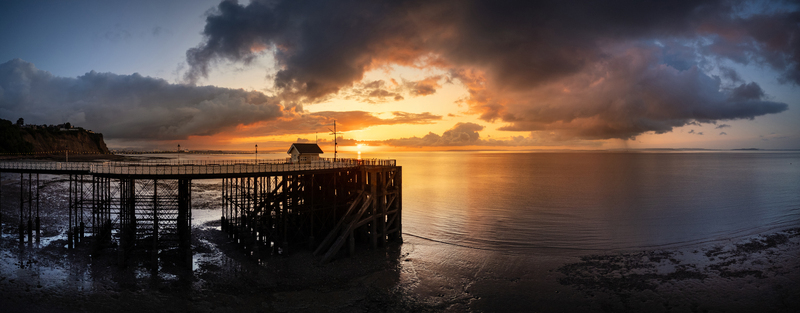 Aerial view of Penarth Pier stretching into the Bristol Channel during a dramatic golden sunrise with dark storm clouds and calm water in Penarth United Kingdom.