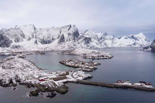 Aerial view of Lofoten featuring snow covered mountains deep blue sea and traditional red fishing huts connected by roads in Nordland Norway.