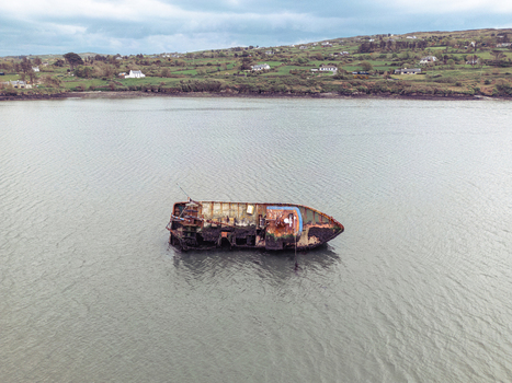 Aerial view of a rusty shipwreck abandoned in shallow coastal waters with green hills and houses in the background under an overcast sky. Aerial view of a rusty shipwreck abandoned in shallow coastal waters with green hills and houses in the background under an overcast sky.
