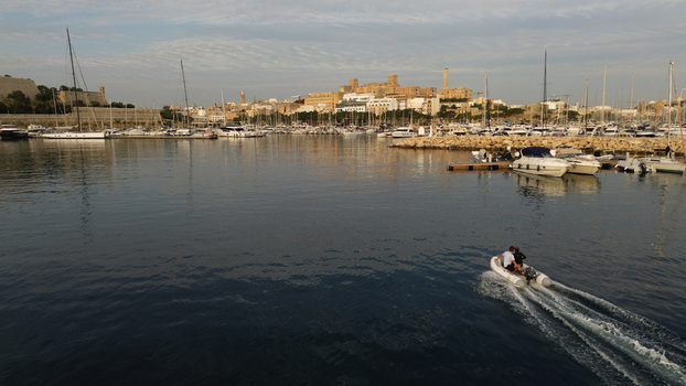Aerial view of a small motorboat cruising through the calm waters of a marina filled with yachts with historic stone buildings and fortifications in the background in Malta. Aerial view of a small motorboat cruising through the calm waters of a marina filled with yachts with historic stone buildings and fortifications in the background in Malta.