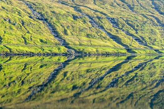 View of the lush green mountainside with cascading waterfalls reflected in the calm water of the fjord Faroe Islands. View of the lush green mountainside with cascading waterfalls reflected in the calm water of the fjord Faroe Islands.