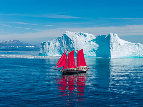 Aerial view of a sailboat with vibrant red sails sailing through deep blue Arctic waters past a massive white iceberg under a clear sky in Greenland. Aerial view of a sailboat with vibrant red sails sailing through deep blue Arctic waters past a massive white iceberg under a clear sky in Greenland.