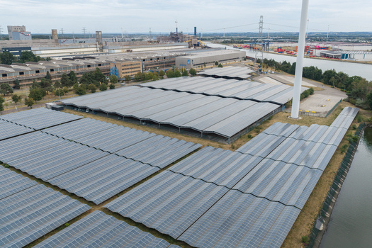Aerial view of a large solar power plant with rows of photovoltaic panels installed at Haven Genk featuring a wind turbine and industrial infrastructure along the canal in Genk Vlaanderen Belgium.