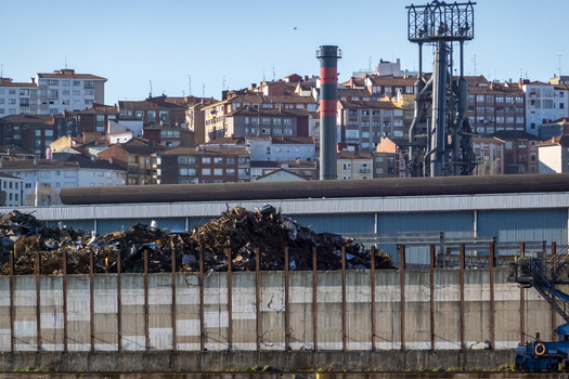 View of a large scrap metal yard with industrial chimneys and residential apartment buildings on a hillside under a clear blue sky in Greater Bilbao Basque Country Spain. View of a large scrap metal yard with industrial chimneys and residential apartment buildings on a hillside under a clear blue sky in Greater Bilbao Basque Country Spain.