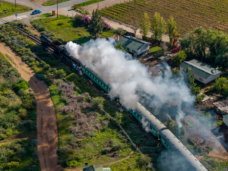 Aerial view of a vintage steam train with white smoke billowing over green carriages through rural vineyards Robertson Western Cape South Africa.