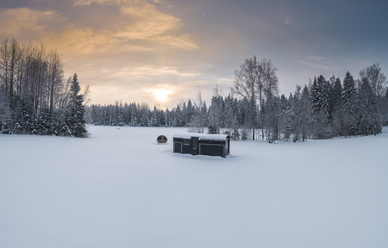 Aerial view of a small dark cabin and a barrel sauna in a vast snow covered field surrounded by a dense forest under a golden sunset sky in Latvia. Aerial view of a small dark cabin and a barrel sauna in a vast snow covered field surrounded by a dense forest under a golden sunset sky in Latvia.