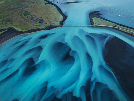 Aerial view of a glacial river delta with turquoise water flowing through black sand sediment and a bridge crossing the landscape in Iceland.