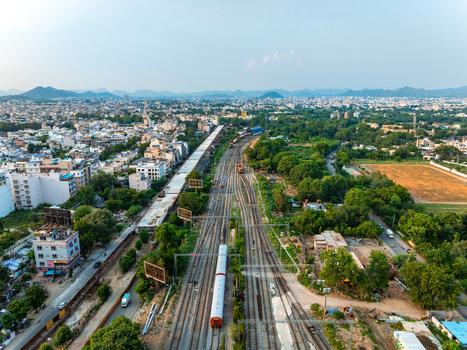 Aerial view of a train moving along the railway tracks through the urban landscape of Udaipur with surrounding buildings lush trees and distant hills in Udaipur Rajasthan India. Aerial view of a train moving along the railway tracks through the urban landscape of Udaipur with surrounding buildings lush trees and distant hills in Udaipur Rajasthan India.