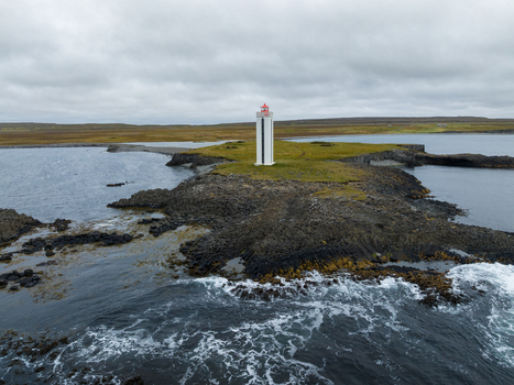Aerial view of a stark white lighthouse with a red top standing tall on a rocky outcrop against the moody sky K&aacute;lfshamarsv&iacute;k H&uacute;nabygg&eth; Iceland.